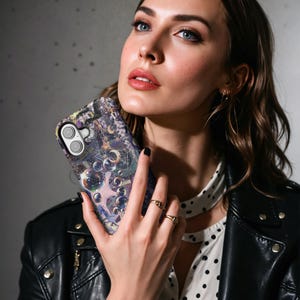 May include: A smartphone case with a celestial design featuring stars, moons, and bubbles in shades of purple, blue, and gold. The case has a glossy finish and a camera cutout. The phone case is held by a person.