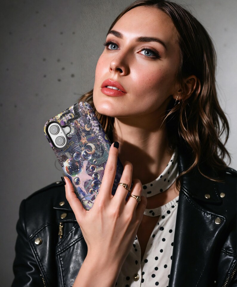 May include: A smartphone case with a celestial design featuring stars, moons, and bubbles in shades of purple, blue, and gold. The case has a glossy finish and a camera cutout. The phone case is held by a person.