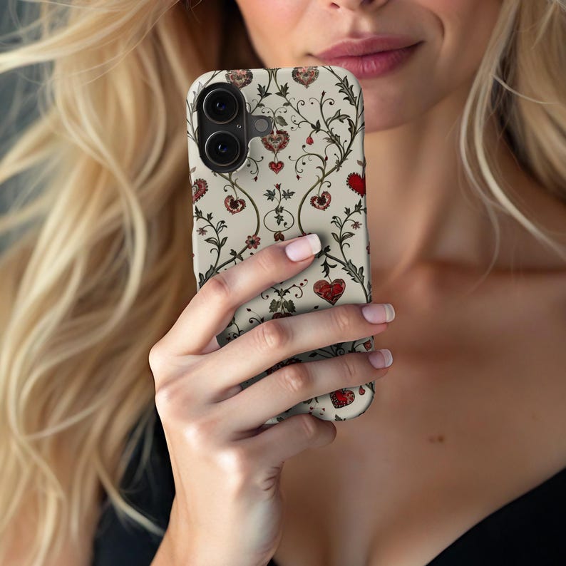 May include: A phone case with a cream background and a repeating pattern of red hearts and floral vines. The case has a dual-lens camera cutout and a smooth, glossy finish. The design is reminiscent of vintage botanical illustrations.