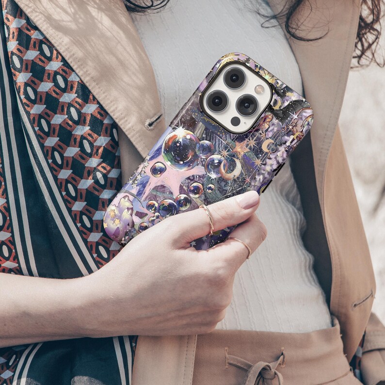 May include: A smartphone case with a colorful, artistic design featuring bubbles and celestial elements. The case has a glossy finish and a protective design, with a camera cutout. The case is held by a person wearing rings.