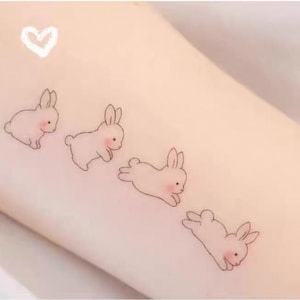May include: Four small, pink bunny tattoos in a row, each in a different pose, with black outlines and rosy cheeks. The bunnies are on a light skin tone, with a white heart and a white shape in the background.