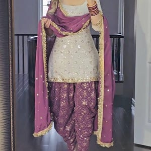 Puede incluir: Un conjunto tradicional indio de salwar kameez. El atuendo presenta una parte superior de color crema con adornos de lentejuelas, combinada con pantalones morados estampados. Una bufanda morada a juego con ribete dorado completa el look. El atuendo se usa con tacones adornados.
