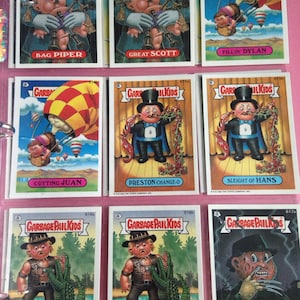 May include: A collection of Garbage Pail Kids trading cards. The cards feature cartoon characters in humorous and grotesque situations. The cards have titles such as "Bag Piper", "Great Scott", and "Fillin' Dylan". The cards are in a pink binder.