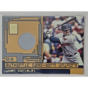 May include: A rectangular sports trading card featuring a photograph of a baseball player swinging a bat. The card includes a circular piece of grey fabric embedded in a gold and grey background with the text AUTHENTIC GAME-WORN UNIFORM JUAN GONZALEZ.