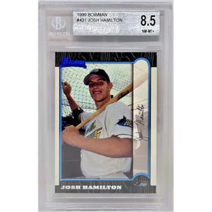 May include: A 1999 Bowman baseball card of Josh Hamilton, graded 8.5 by Beckett. The card features a color image of Hamilton in a baseball uniform holding a bat. The card is encased in a clear protective holder.