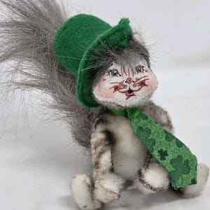 May include: A plush squirrel doll with a green hat and scarf, designed with a St. Patrick's Day theme. The squirrel has a gray and white body, a fluffy gray tail, and a painted face with blue eyes and pink cheeks. The green scarf features a shamrock pattern.