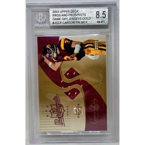 以下が含まれることがあります： カーソン・パーマーをフィーチャーした、グレーディングされた2003 Upper Deck Pros and Prospects Game Day Jerseys Goldトレーディングカード。カードはベケットホルダーに収められ、8.5のグレードです。カードは、マルーンとゴールドのユニフォームを着たフットボール選手を背景に金色の背景を持っています。