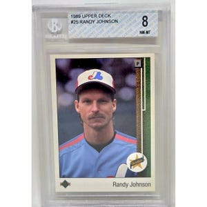 Puede incluir: Una tarjeta de béisbol Upper Deck de 1989 de Randy Johnson, calificada NM-MT 8 por Beckett. La tarjeta presenta un retrato de Johnson con una camiseta de béisbol azul y una gorra con un logotipo. La tarjeta está en un soporte protector.