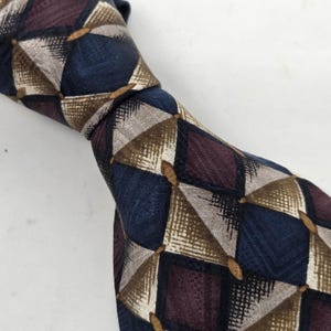 Puede incluir: Primer plano de una corbata estampada. La corbata presenta un diseño geométrico repetitivo de cuadrados y triángulos en azul marino, burdeos, beige y gris. La corbata está anudada, mostrando la textura y el patrón de la tela.