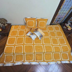 May include: A handmade mustard yellow crochet blanket with a square pattern, paired with matching square pillows. The blanket and pillows have white accents. Decorative pillows in shades of gold and silver complete the bed set.