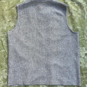 Pu&ograve; includere: Un gilet grigio senza maniche realizzato in tessuto strutturato. Il gilet ha uno scollo a V e una vestibilit&agrave; su misura. Il capo &egrave; esposto su un tessuto verde a fantasia floreale.