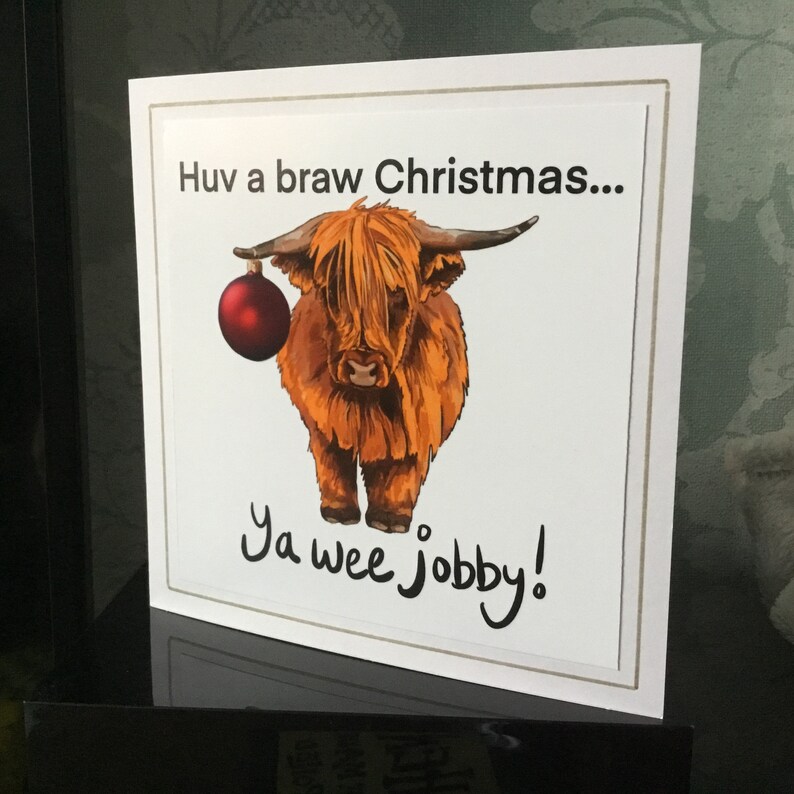 Puede incluir: Una tarjeta de Navidad con una vaca de las Tierras Altas de Escocia de dibujos animados con un adorno rojo. La tarjeta dice "Huv a braw Christmas... Ya wee jobby!"