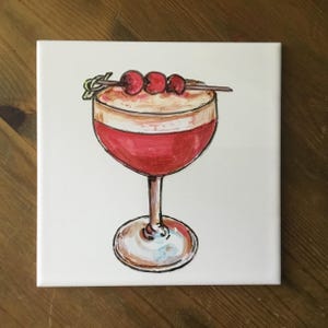 Op de afbeelding: Een handgeschilderde keramische tegel met een rode cocktail met een witte schuimlaag en drie rode kersen aan een spies. De cocktail zit in een glas met een steel en een heldere bodem.