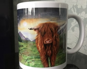 May include: A white ceramic mug with a colorful watercolor illustration of a Highland cow in a field. The cow is brown and has long, shaggy hair. The background is a watercolor painting of a mountain range and a sunset.