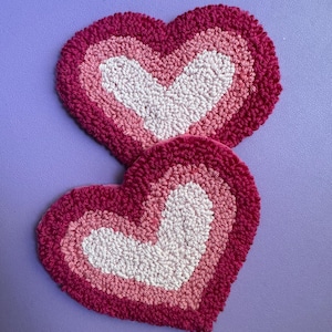 May include: Two heart-shaped coasters, handmade with a looped yarn. The coasters feature a layered design in shades of pink and white. The gradient effect is visible in the design. The coasters are placed on a purple surface.