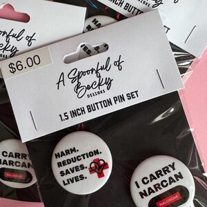 May include: A set of two white 3.8 cm button pins on a display card labelled A Spoonful of Becky Designs. One pin reads HARM. REDUCTION. SAVES. LIVES. with a red medical cross and heart icon. The other pin reads I CARRY NARCAN with a red NALOXONE label.