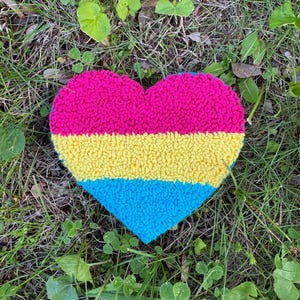 May include: A heart-shaped rug with a textured finish, featuring three horizontal stripes. The colours are hot pink, yellow, and turquoise blue. The rug is placed on grass and clover, creating a natural backdrop.