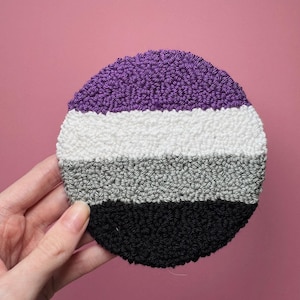 Peut inclure: Un dessous de verre circulaire en punch needle composé de quatre bandes horizontales violettes, blanches, grises et noires. Cette pièce textile présente une surface en fil bouclé texturé et une forme ronde uniforme avec un design coloré.