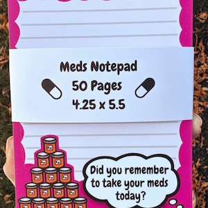 Peut inclure: Un bloc-notes rose avec des pages lignées et une bande blanche avec le texte "Meds Notepad, 50 Pages, 10.8 x 14 cm". Le bloc-notes a une bordure décorative et une bulle de dialogue qui dit "Did you remember to take your meds today?"