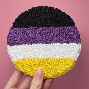 Peut inclure: Un dessous de verre circulaire en punch needle avec des rayures horizontales noires, violettes, blanches et jaunes. La surface textile présente une texture en bouclettes, formant un motif distinct de rangées de fils colorés en cercle.