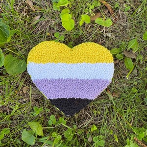 Peut inclure: Un tapis en forme de cœur avec le drapeau de la fierté non binaire. Le tapis présente des rayures horizontales jaunes, bleu clair, lavande et noires. Il est posé sur de l'herbe et du trèfle. Fait à la main.