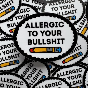 May include: A pile of white, circular stickers with a black, scalloped edge. Each sticker displays the text "ALLERGIC TO YOUR BULLSHIT" in bold black lettering, accompanied by a pencil illustration. The stickers are arranged randomly across the image.