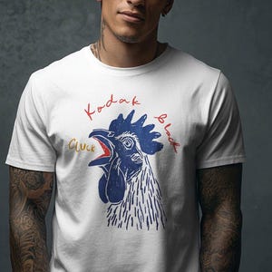 Puede incluir: Camiseta blanca con un gráfico de gallo azul y rojo y las palabras "Kodak Black" y "Cluck". El gallo tiene el pico abierto y una cresta detallada. El texto es en rojo y azul.