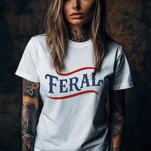 Può includere: T-shirt bianca con la scritta "FERAL" in blu navy, incorniciata da un banner rosso. La maglietta a maniche corte sembra essere di cotone. L'immagine mostra la maglietta su una persona tatuata.