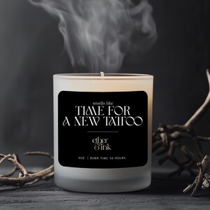 Puede incluir: Una vela de vidrio esmerilado con una etiqueta negra que dice "smells like TIME FOR A NEW TATTOO" y "ether & ink". La vela es de 255g y tiene un tiempo de combustión de 50 horas. Volutas de humo se elevan de la mecha encendida.