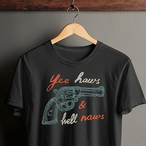Może przedstawiać: Czarny t-shirt z grafiką rewolweru i napisem "Yee haws & hell naws" w kolorze pomarańczowym i białym. Rewolwer jest obrysowany na niebiesko. Koszulka wisi na drewnianym wieszaku.