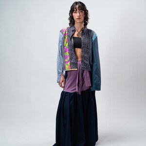 Puede incluir: Una chaqueta patchwork con varios patrones de tela, incluyendo diseños florales y a rayas, se lleva sobre un top negro. Una falda larga azul oscuro completa el conjunto. La modelo lleva un collar.