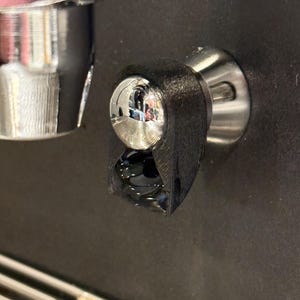 May include: Close-up of a black plastic coffee machine part with a silver metal knob. The part is attached to a black surface.