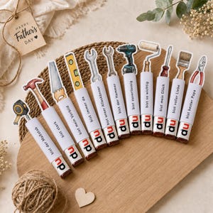 Pode incluir: Uma cole&ccedil;&atilde;o de barras de chocolate com coberturas tem&aacute;ticas de ferramentas, dispostas em uma t&aacute;bua de madeira. Cada barra tem uma ilustra&ccedil;&atilde;o de ferramenta diferente e uma frase em alem&atilde;o. Uma etiqueta com "Happy Father's Day" est&aacute; anexada.