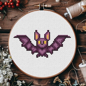 May include: A cross-stitch embroidery of a purple bat with yellow eyes and ears, set in a wooden hoop. The bat design is on white fabric. The background is a wooden surface with decorative elements.