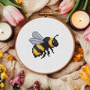 May include: A cross-stitch embroidery hoop with a white background and a yellow and black bee design. The bee has grey wings and is stitched in a pixelated style.