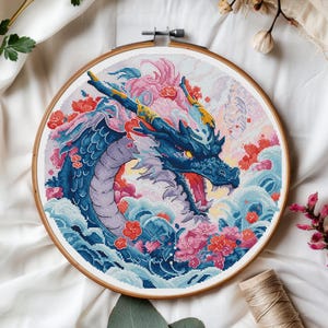 Könnte beinhalten: Ein runder Stickrahmen mit einem farbenfrohen Kreuzstich-Design eines blauen Drachen mit rosa und weißen Blumen. Der Drache ist von blauen und weißen Wellen umgeben.