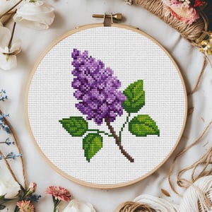 May include: A purple lilac flower with green leaves is embroidered on white fabric in a round wooden embroidery hoop.