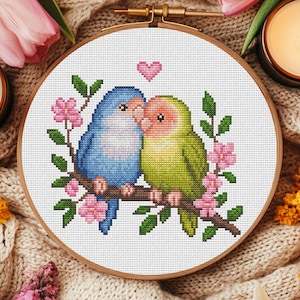 May include: A cross-stitch embroidery in a round wooden hoop featuring a blue bird and a green bird perched together on a brown branch. The birds are surrounded by pink flowers and green leaves, with a small pink heart stitched above them.