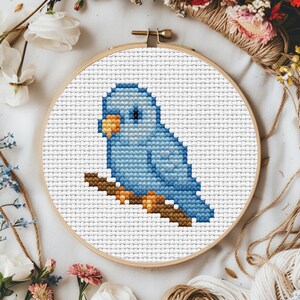 May include: A cross-stitch embroidery of a blue bird perched on a brown branch, set within a wooden hoop. The bird has a yellow beak and black eye. The background is white fabric, and the hoop is surrounded by flowers and twine.