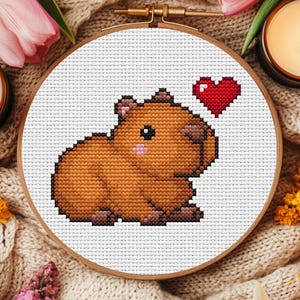 May include: A cross-stitch embroidery of a capybara with a red heart above its head, set in a wooden hoop. The capybara is brown with pink accents. The background is white, and the hoop is surrounded by a cozy, neutral-toned fabric.