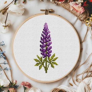 Może przedstawiać: Wzór haftu krzyżykowego przedstawiający fioletową kwiat lupiny z zielonymi liśćmi na białym tle. Wzór jest oprawiony w drewniany tamborek do haftu.