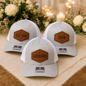 May include: Three grey and white baseball caps with mesh backs. Each cap features a brown hexagonal leather patch on the front. The patches are engraved with the text BEST MAN, GROOM, and GROOMSMEN, along with the date 5.21.24 on each.