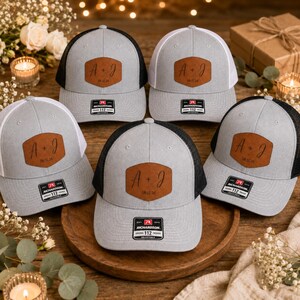 May include: Five trucker hats featuring a light gray front panel and a brown leather patch with the text A + J and 08.12.26. Two hats have black mesh backs, while three hats have white mesh backs. Each hat includes a black Richardson 112 sticker.