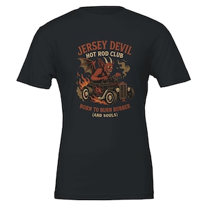 Puede incluir: Camiseta negra con un gráfico de un diablo rojo conduciendo un hot rod, con llamas y humo. El texto dice "Jersey Devil Hot Rod Club" y "Born to Burn Rubber (and Souls)". El diseño es en tonos de rojo, naranja y marrón.