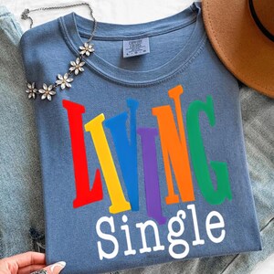 K&ouml;nnte beinhalten: Ein blaues T-Shirt mit den Worten "LIVING SINGLE" in bunten Blockbuchstaben. Das Shirt hat einen Rundhalsausschnitt und kurze &Auml;rmel. Eine Blumenkette und ein brauner Hut sind ebenfalls zu sehen.