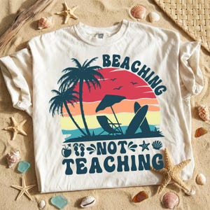 May include: Cream-colored t-shirt with a beach-themed graphic. The design features palm trees, a sunset, a beach umbrella, a surfboard, and the text "BEACHING NOT TEACHING" in a retro font. The shirt is displayed on a sandy surface with seashells.
