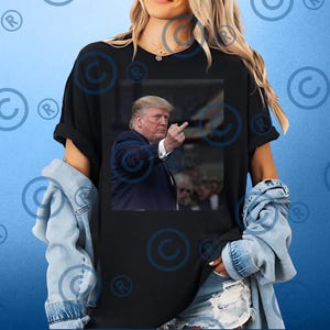 May include: Black t-shirt featuring a photographic image of a man in a suit, raising his middle finger. The image is centered on the shirt. The person wearing the shirt is also wearing a denim jacket.