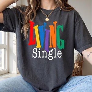 K&ouml;nnte beinhalten: Dunkelgraues T-Shirt mit dem Schriftzug "LIVING SINGLE" in bunten, fetten Buchstaben. Das Wort "LIVING" ist in Rot, Gelb, Blau, Lila, Orange und Gr&uuml;n gehalten. Das Wort "SINGLE" ist wei&szlig;. Ein l&auml;ssiger Unisex-Stil.