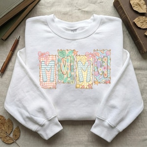 May include: White sweatshirt with the word "MAMA" in colorful, decorative letters. Each letter is designed with a different pattern, including stripes, tropical leaves, and seashells. The sweatshirt has a crew neck and long sleeves.