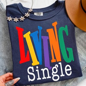K&ouml;nnte beinhalten: Marineblaues Rundhals-T-Shirt mit den Worten "LIVING Single" in bunten, fetten Buchstaben. Das Wort "LIVING" ist in Rot, Gelb, Blau, Lila, Orange und Gr&uuml;n gehalten. Das Wort "Single" ist in Wei&szlig; gehalten. Eine Blumenkette liegt auf dem Shirt.